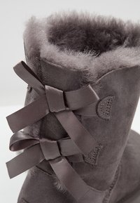 Szare zamszowe botki z futrzanym wykończeniem, posiadające ozdobne szare wstążki związane z tyłu i przeszywane detale po bokach.