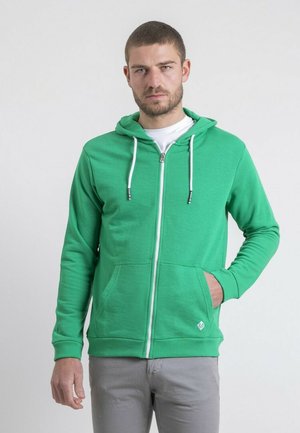 Un homme aux cheveux courts et à la barbe, portant un sweat à capuche vert zippé avec des cordons blancs et un pantalon gris, debout avec une main dans la poche.