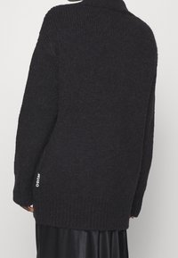 Schwarze Strickjacke mit hohem Kragen, langen Ärmeln und geripptem Saum. Mit einem gewebten Logo-Detail auf der unteren linken Seite.