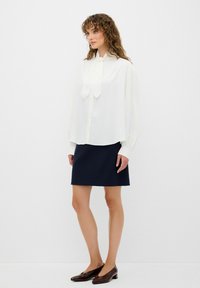 Blouse blanche avec un col à nœud, devant boutonné et manches longues. Associée à une mini-jupe marine et des chaussures à talons brunes. Design simple.