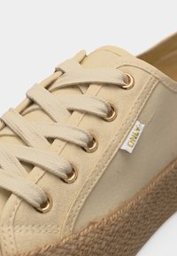 Μπεζ αθλητικό παπούτσι από καμβά με κρεμ κορδόνια, χρυσά μεταλλικά τρύπες, σόλα πλατφόρμα από υφαντό γιούτα και μικρή λευκή ετικέτα στο πλάι.