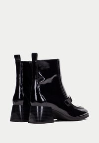 Botas de tobillo de charol negro con tacón cuadrado, cierre de cremallera y detalle de correa decorativa en el lateral. Textura suave y brillante.
