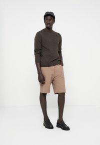 Brunt stickad tröja, beige texturerade shorts och svarta slip-on skor, modellen står mot en enkel vit bakgrund, avslappnad outfit avbildad.