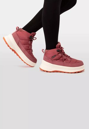 Personne portant des baskets à plateforme de hauteur moyenne couleur grenat avec semelles blanches et reliefs orange, et des leggings noirs sur un fond neutre.