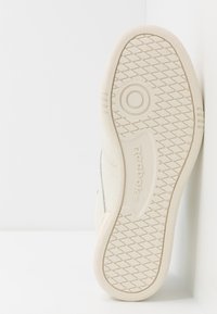 Suela de zapatilla Reebok en goma de color blanco roto, con un emblema circular, patrón de diamante en la suela y logotipo grabado en el talón.
