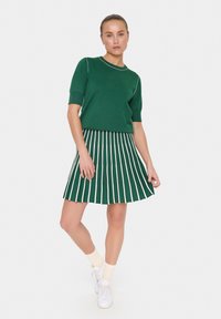 Haut en tricot vert à manches courtes avec des bordures blanches, associé à une jupe plissée verte avec des rayures verticales blanches. Baskets blanches et chaussettes claires.