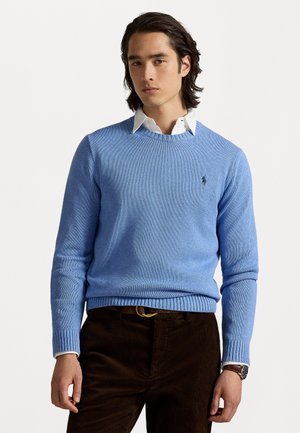 Light blue sweater with a textured knit, round neckline, and ribbed cuffs, paired with a white collared shirt and brown corduroy pants.