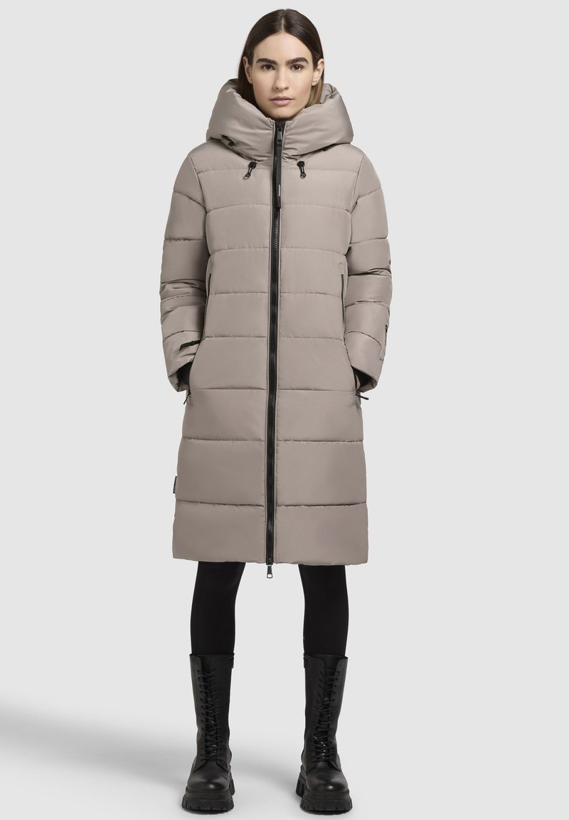 Beige steppelt kabát kapucnival, középen cipzárral, steppelt részekkel és oldalzsebekkel. Fekete leggings és fűzős fekete csizmák fölött viselve.