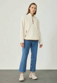 Cremfarbener Fleecepullover mit halbem Reißverschluss-Kragen, braunen Akzenten und seitlichen Taschen, kombiniert mit blauen Straight-Leg-Jeans und beigen Sneakers.
