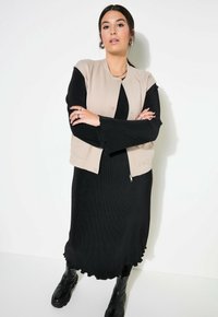 Beige ärmelloser Weste über einem schwarzen, gerippten langen Kleid mit geschwungener Saumlinie. Das Modell trägt schwarze kniehohe Stiefel und geschichteten Schmuck.