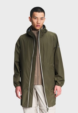 Hombre con un impermeable verde oliva con capucha sobre una chaqueta marrón claro con cremallera y pantalones cargo beige, de pie frente a un fondo liso.