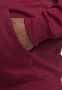 Mano de piel oscura con un tatuaje de cruz descansando dentro del bolsillo delantero de un sudadera color burdeos con mangas de puño acanalado.