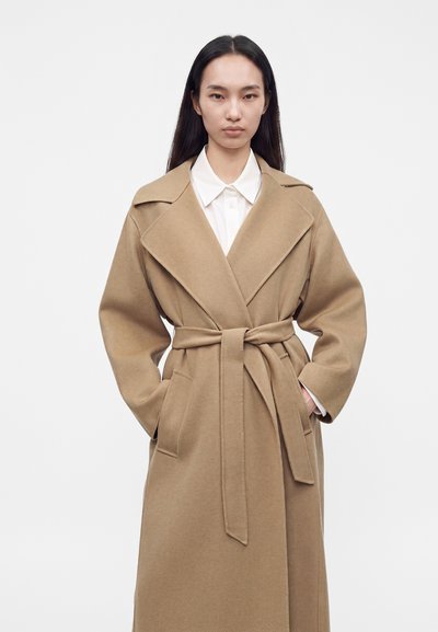 Femme portant un manteau en laine beige ceinturé par-dessus une chemise blanche à col, debout les mains dans les poches sur un fond clair uni.