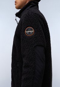 Giacca in pile nera con un'esterno testurizzato, colletto alto e tasche con zip. Include una spilla rotonda con il logo sul braccio sinistro.