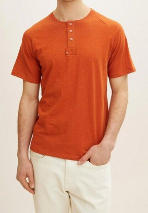 T-shirt à manches courtes et col rond de couleur orange profond avec une patte de boutonnage et un tissu texturé. Associé à un pantalon de couleur claire.