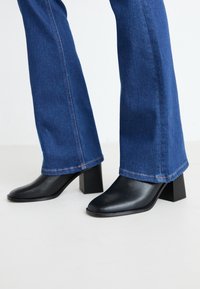 Jean en denim bleu porté avec des bottines à talons noires sur un fond clair, montrant les jambes inférieures et les détails des chaussures.