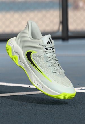 Zapatilla deportiva gris claro con el logo de Nike en negro y amarillo neón, parte superior de malla detallada y suela amarilla neón sobre una superficie de cancha azul.