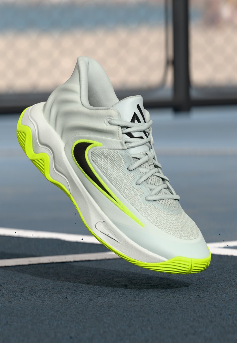 Lys grå atletisk sko med sort og neongul Nike swoosh, detaljeret mesh-overdel og neongul sål på en blå baneflade.