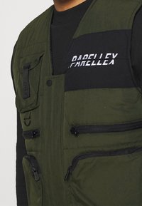 Grüne gesteppte Weste mit schwarzen Akzenten, mehreren Reißverschlusstaschen und gesticktem "PARELLEX"-Logo auf der Brust. Strukturierter Stoff.