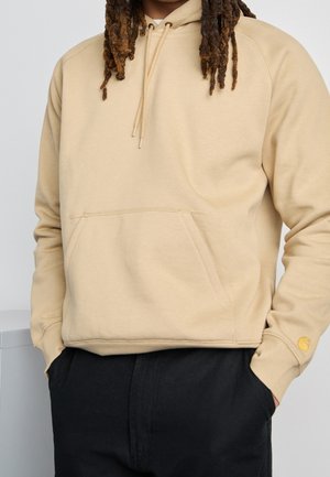 Sudadera con capucha beige con bolsillo delantero y cordones, llevada por una persona con rastas y pantalones negros, con las manos en los bolsillos.