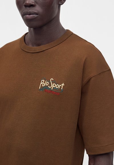 Άνδρας που φοράει καφέ μπλουζάκι Polo Sport Sportsman με κεντημένο λογότυπο στο στήθος, εμφανίζεται από τους ώμους μέχρι τη μέση του κορμού σε απλό φόντο.