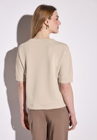 Béžový top s krátkým rukávem, uvolněným střihem, kulatým výstřihem a hladkou texturou. Spárován s elegantními hnědými kalhotami. Minimalistický design.
