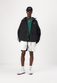 Corta-vento preto com riscas brancas, camisa verde, calções brancos e sapatos cinzentos. A jaqueta apresenta grandes bolsos frontais e um capuz.