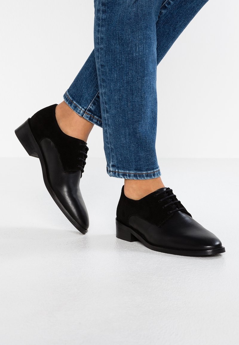 Chaussures noires en cuir lisse et en daim. Bout arrondi, six œillets et un petit talon empilé. Portées avec un jean bleu.