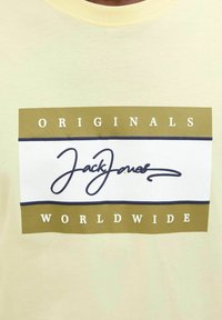 Lys gul kortærmet t-shirt med et rektangulært grafik, der har "JackJones" i blå kursiv, omgivet af teksten "ORIGINALS" og "WORLDWIDE".