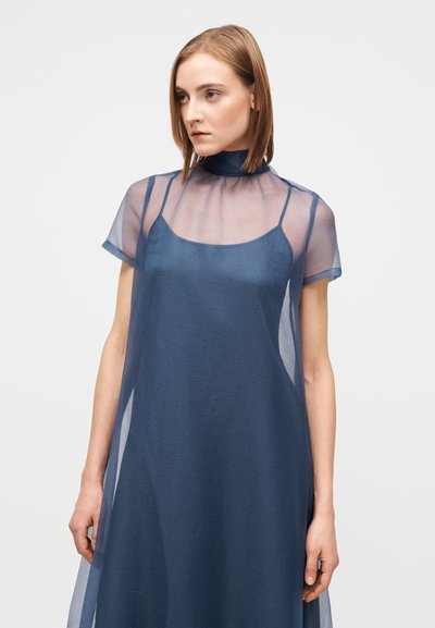 Γυναίκα με μαλλιά μέχρι τον ώμο, ντυμένη με διάφανο φόρεμα ναυτικού μπλε κοντού μανικιού πάνω από αμάνικο φόρεμα-φόρεμα σε ναυτικό μπλε, κοιτάζει προς τα αριστερά.