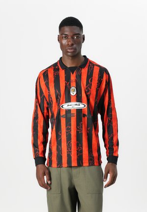 Maglia da calcio a maniche lunghe con audaci righe verticali nere e rosse, colletto nero e logo centrale su sfondo bianco.