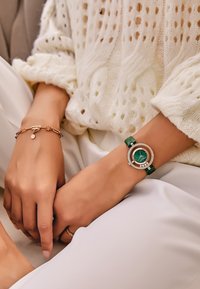 Reloj verde con esfera redonda, acentos plateados y una correa a juego. Acompañado de una pulsera de oro rosa con un charm circular. Suéter de punto blanco texturizado.