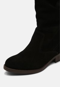 Bottine en daim noir avec bout arrondi, couture visible et semelle à talon bas de couleur marron.