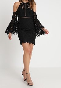 Robe en dentelle noire avec des manches évasées dénudées, un corps ajusté, un motif floral et une finition texturée. Portée avec des talons noirs à lanières.