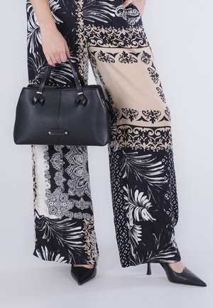 Vrouw met zwarte leren handtas, draagt wijde broek met zwart, beige en wit ingewikkeld bloemen- en geometrisch patroon en zwarte hakken.