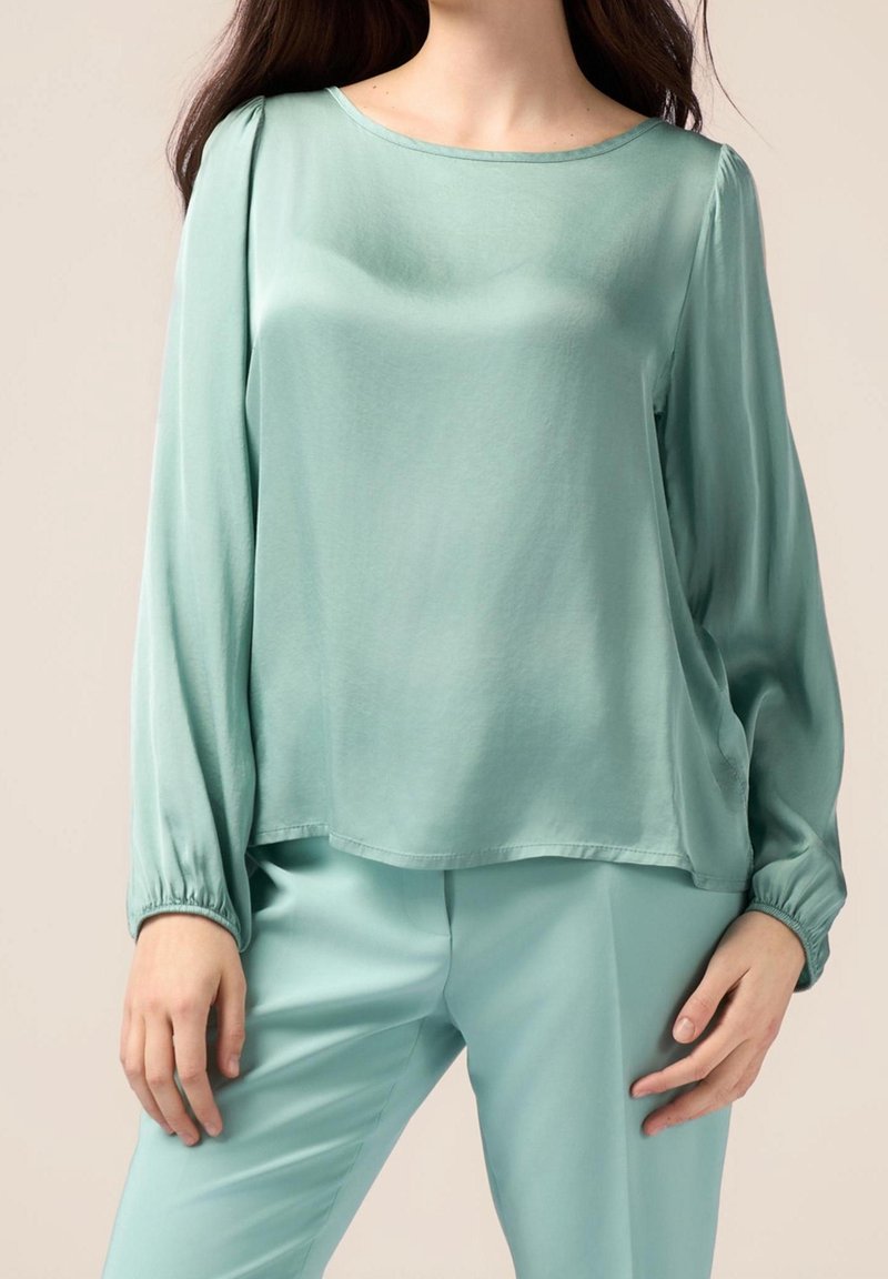 Femme portant un chemisier en satin vert clair à manches longues avec un pantalon vert clair assorti, debout devant un fond uni.