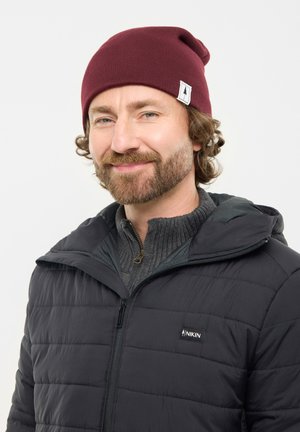 Smiling man with beard wearing a maroon beanie and black puffer jacket over a gray zip-up sweater against white background.