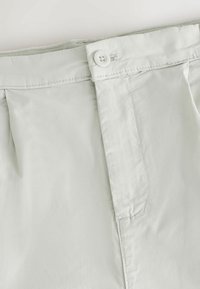 Gros plan d’un pantalon gris clair montrant la ceinture avec un unique bouton blanc et un détail de braguette cousue.