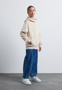 Hanorac bej cu glugă, croială oversized, din amestec de bumbac. Blugi albaștri, largi. Sneakerși cu accente pastel și talpă albă.
