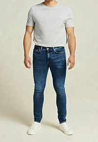 Man med en enkel vit t-shirt med kort ärm, mörkblå skinny jeans och vita sneakers som står mot en neutral bakgrund.