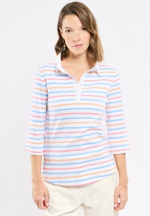 Femme portant une chemise à col avec des rayures pastel horizontales en bleu, rose et orange, associée à un pantalon de couleur claire et des boucles d'oreilles dorées.