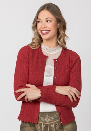 Lächelnde Frau mit welligem Haar trägt eine rote Strickjacke über einem weißen Spitzenoberteil, verschränkt die Arme und schaut seitlich vor schlichtem Hintergrund.