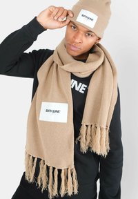 Beige gebreide sjaal met franjes aan de uiteinden, met een wit label waarop 'SIXTH JUNE' staat. Draag het met een bijpassende beige beanie en een zwart longsleeve shirt.