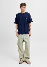 Granatowa bawełniana koszulka z krótkim rękawem, biała logo na piersi; jasnoszare szerokie dżinsy; beżowe buty.