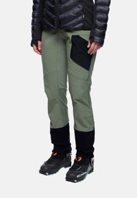 Groene softshell broek met kniestukken, een zwarte zijzak en zwarte manchetten. Draagt zwarte laarzen met oranje accenten en lichtblauwe zolen.