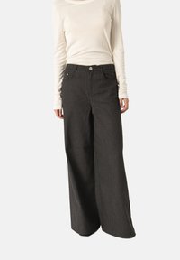 Celana panjang lebar berwarna abu-abu gelap dengan tekstur halus, dilengkapi dengan saku depan dan penutupan kancing, dipadukan dengan atasan lengan panjang berwarna beige muda.