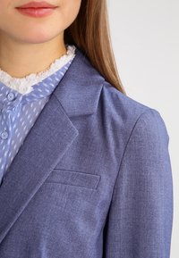 Jasnoniebieska marynarka z klapami i kieszonką na piersi, wykonana z teksturowanej tkaniny. Nałożona na jasnoniebieską koszulę w kropki.