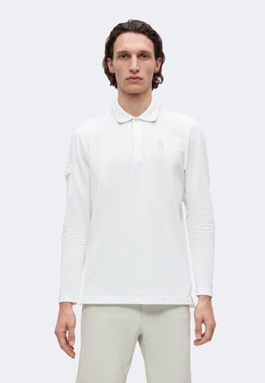 Homme aux cheveux bruns mi-longs porte un polo blanc à manches longues et un pantalon clair, debout devant un fond uni et clair.