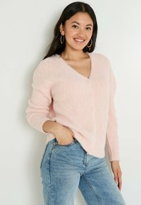 Cardigan en peluche rose clair avec un col en V, motif texturé et fermeture à boutons. Associé à un jean en denim bleu.