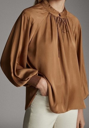 Blouse longue en satin marron à manches longues avec col à volants froncés, col noué et poignets smockés, portée avec un pantalon beige clair.